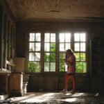 A woman standing alone in an empty, sunlit room, deep in thought — a moment of quiet healing for women rebuilding their lives in silence.