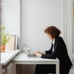 Woman in a black blazer working on laptop at home office, starting before she feels ready to build her future self.