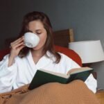 A woman in a white robe sipping from a cup while reading a book under a blanket, embodying a calm and intentional morning routine focused on self-care and mindfulness.